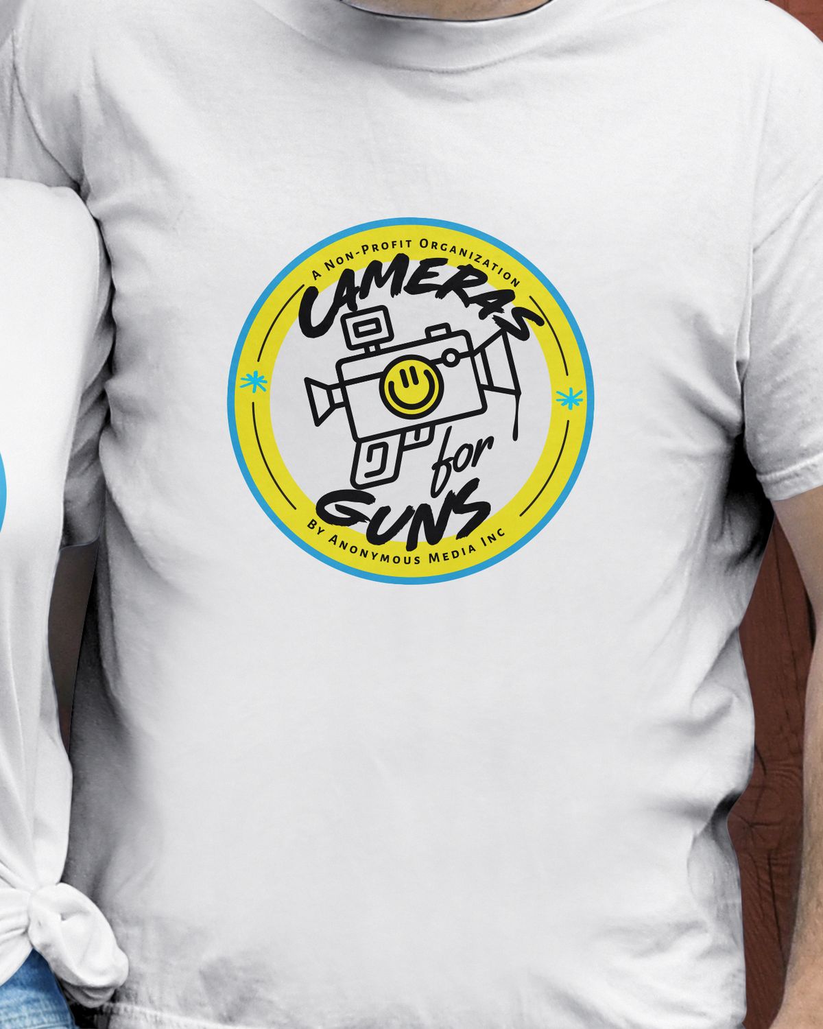 White unisex t-shirt with the Cameras for Guns badge on the chest, worn against a red wooden wall